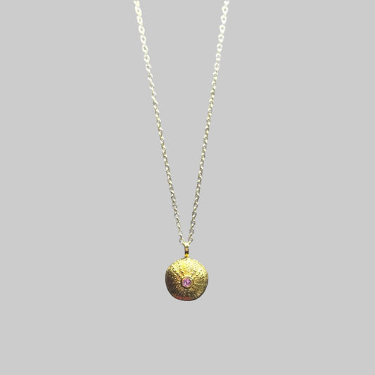 Gold necklace with a round pendant featuring a pink gemstone on a light gray background