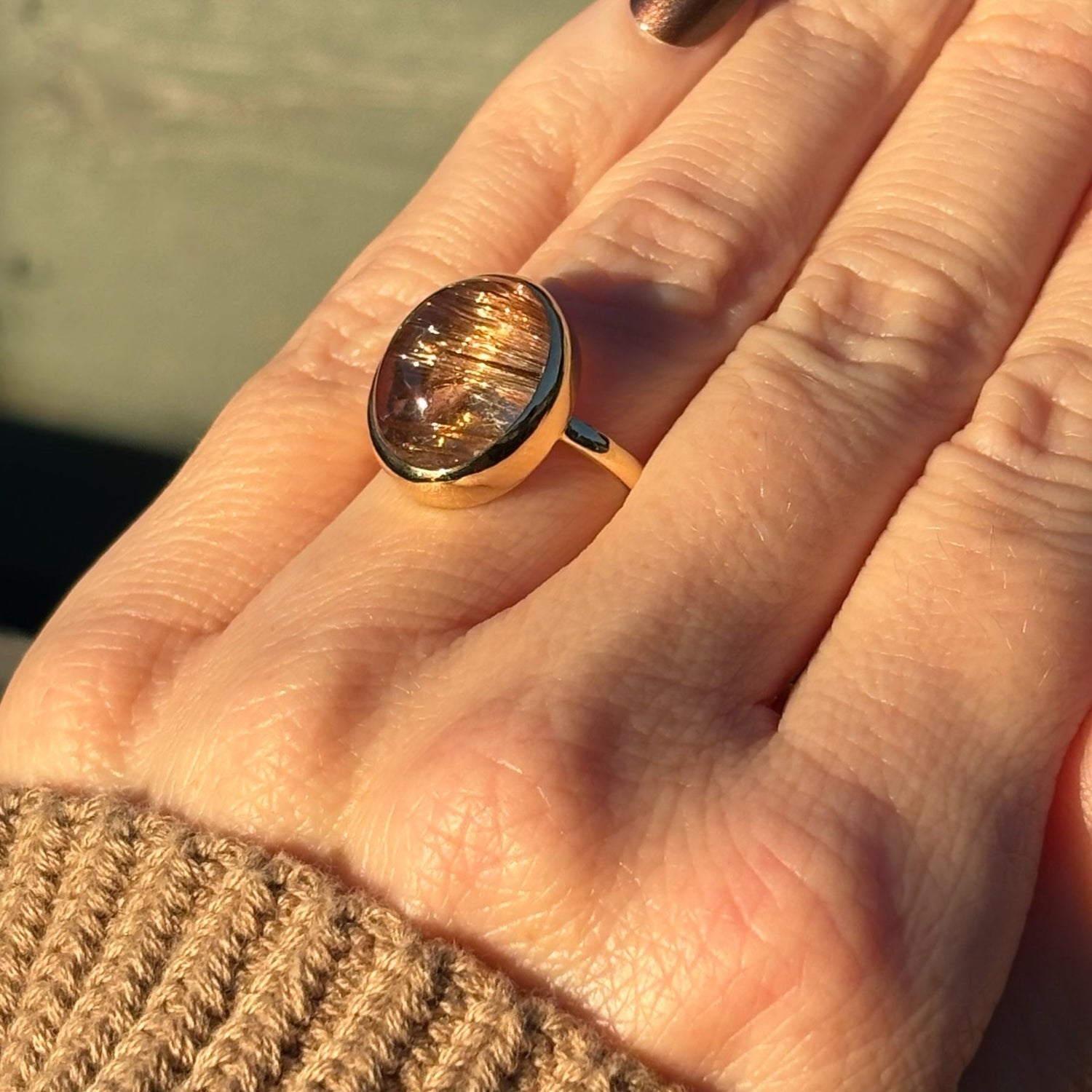 Close-up of a hand wearing a gold ring with a large gemstone, against a blurred background.