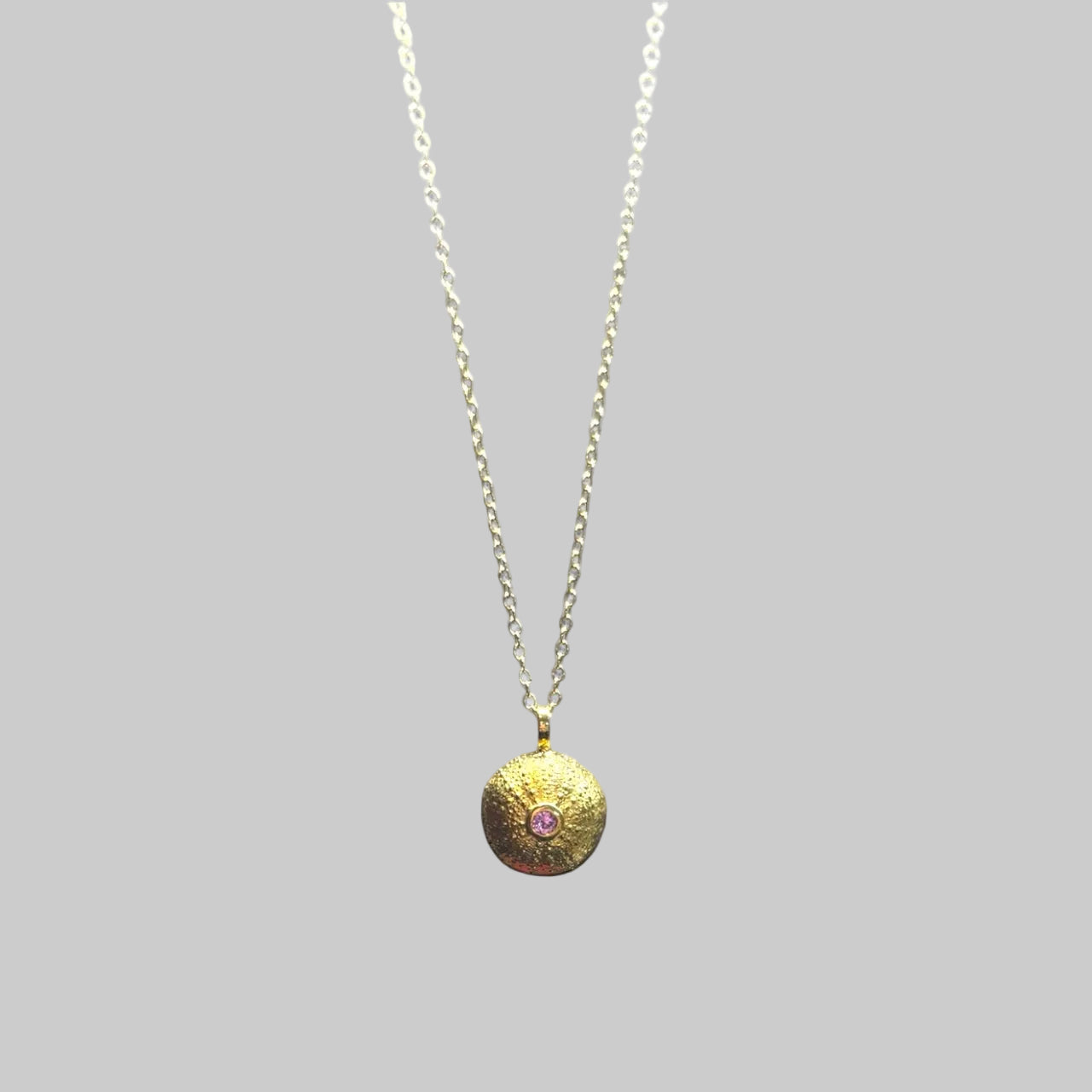 Gold necklace with a round pendant featuring a pink gemstone on a light gray background